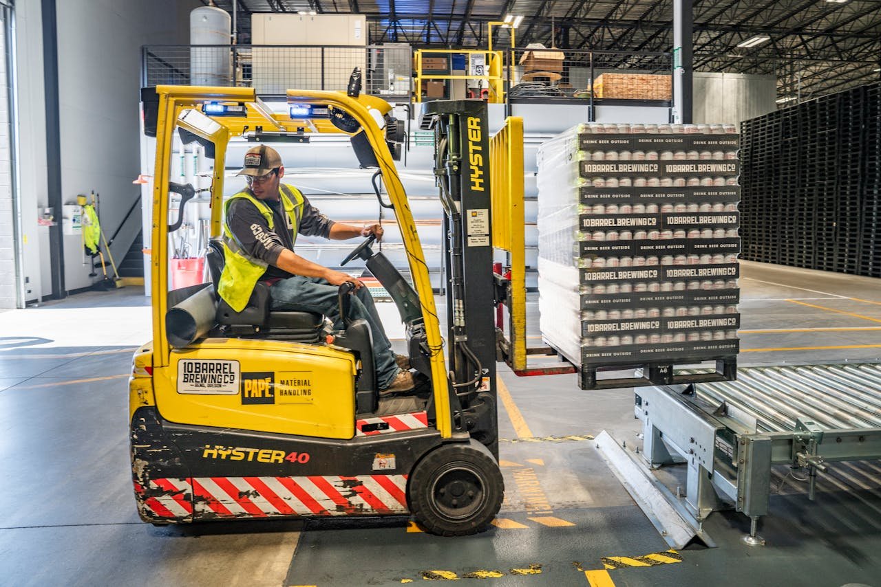 why-choose-us A warehouse worker maneuvers a forklift to transport crates for brewing company storage.