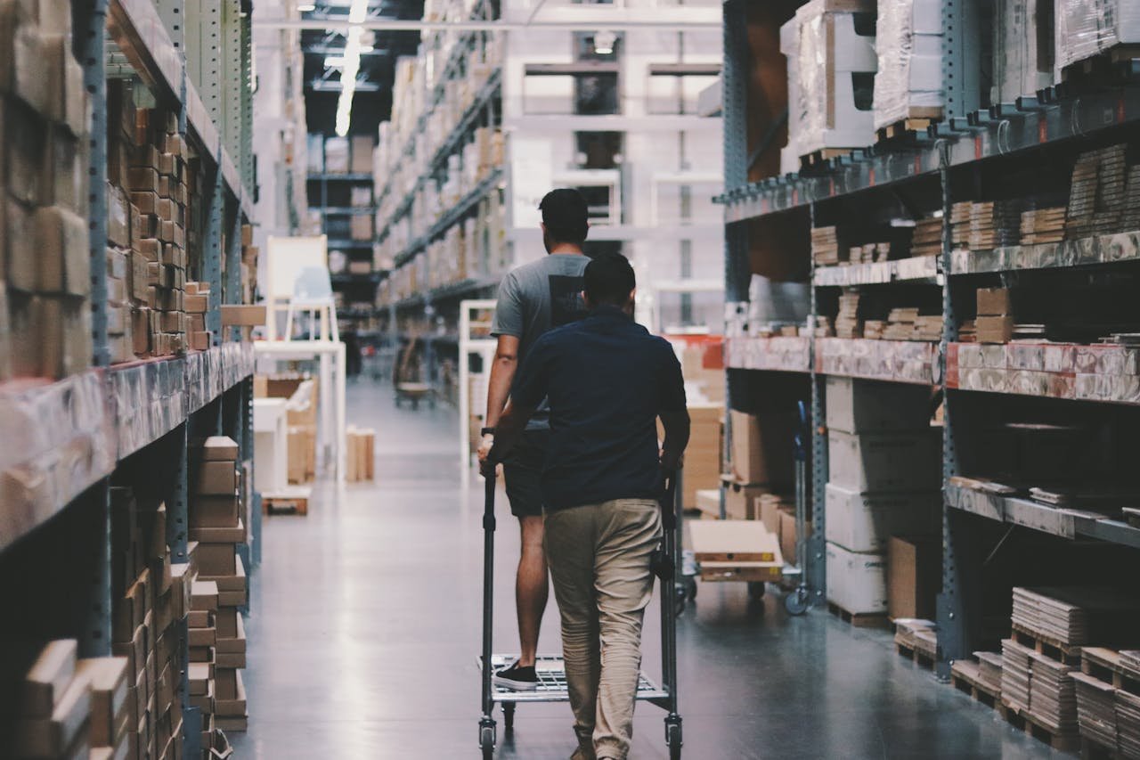 our-journey-02 Two men maneuver a trolley in a large warehouse filled with boxes and shelves.